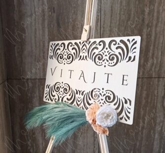Drevená uvítacia tabuľa s gravírovaným nápisom ‚Vitajte na našej svadbe‘, zdobená kvetmi a pampovou trávou – elegantná svadobná výzdoba.
