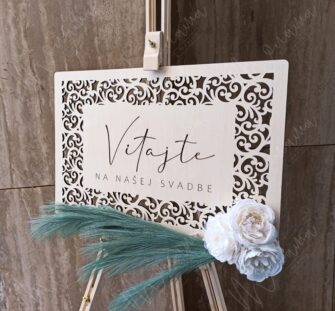 Drevená uvítacia tabuľa s gravírovaným nápisom ‚Vitajte na našej svadbe‘, zdobená kvetmi a pampovou trávou – elegantná svadobná výzdoba.