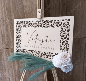 Drevená uvítacia tabuľa s gravírovaným nápisom ‚Vitajte na našej svadbe‘, zdobená kvetmi a pampovou trávou – elegantná svadobná výzdoba.