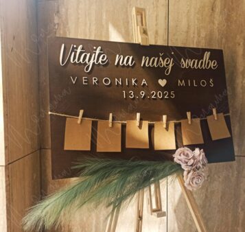 Drevený zasadací poriadok na svadbu – tmavá tabuľa s nápisom Vitajte na našej svadbe, menami, dátumom a šnúrkou so štipcami na kartičky hostí