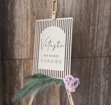 Uvítacia tabuľa na svadbu – elegantná drevená dekorácia s nápisom Vitajte na našej svadbe, doplnená o kvety a prírodné trávy.